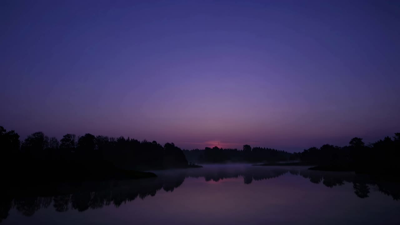 A serene twilight landscape video with a wide-angle view captures a calm lake reflecting the purple