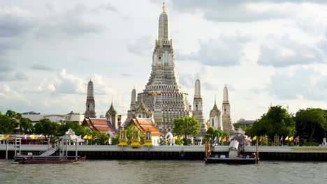 shot of beautiful wat arun temple in bangkok, thailand with the view of motor boats passing by carrying tourists during evening time