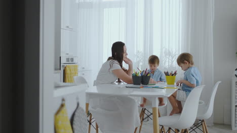 Mom-helps-sons-learn-to-draw-doing-homework-preschool-preparation-at-home-sitting-in-the-white-kitchen.-Two-brothers-draw-a-portrait-of-mom-together