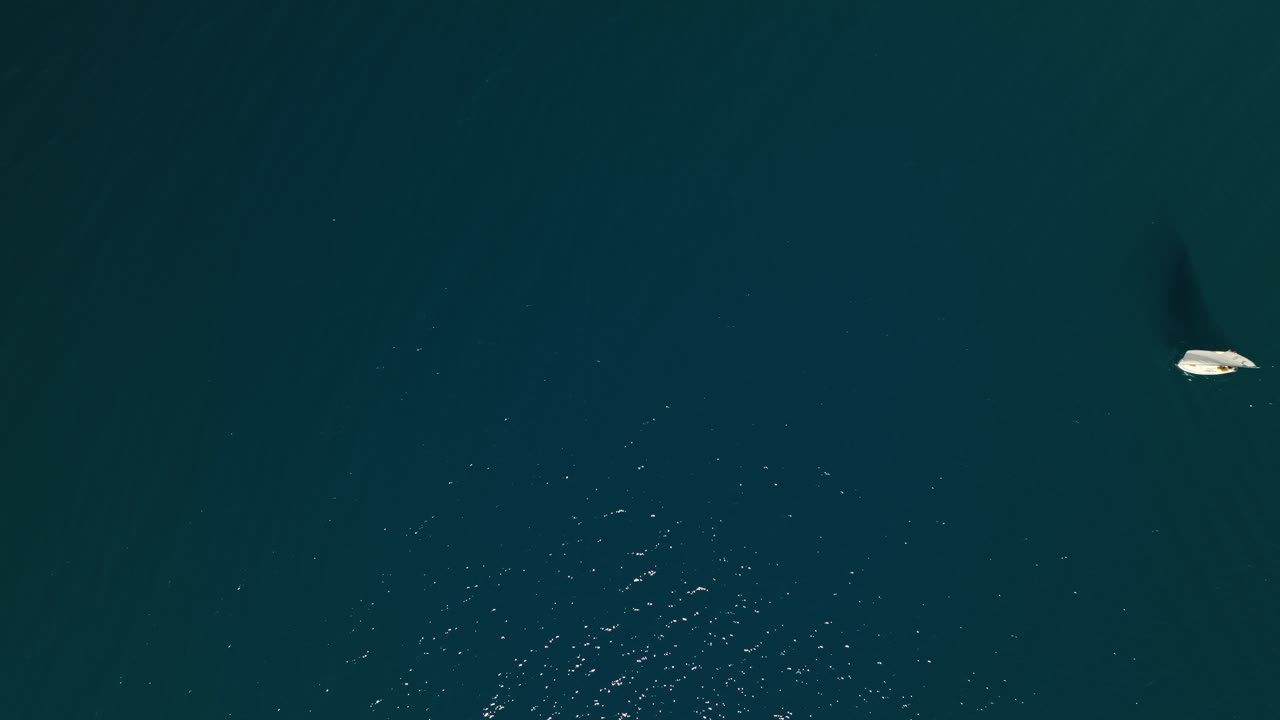 Drone top down rises as sailboat with sail shadow enters frame floating