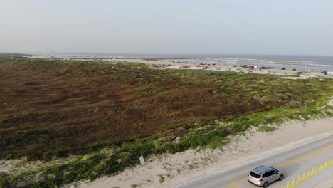 Antena-Decente-Desde-La-Vista-De-La-Playa-Y-Los-Pastizales,-Aterrizando-En-El-Estacionamiento-Con-2-Vehículos-Pasando