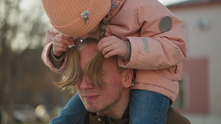 A little girl in a pink cap and jacket sits joyfully on her dad's neck as they walk through a sunlit park. She playfully bends her head down and uses her hair to cover his eyes