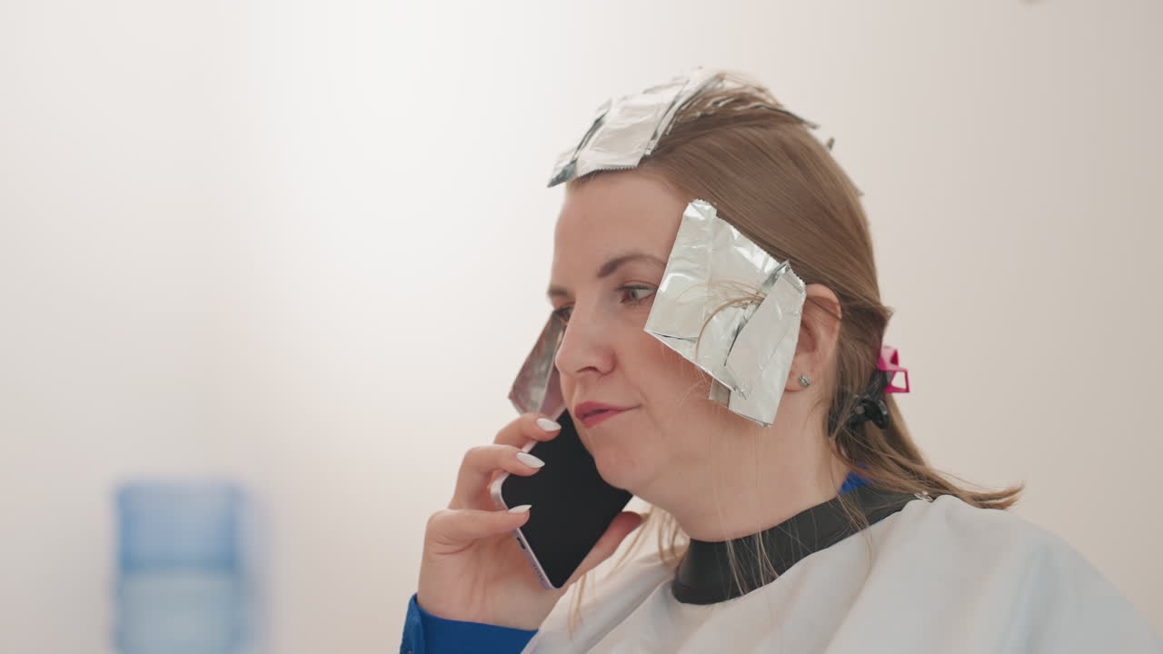 white woman in salon with foils talking on phone, wearing cape while highlights process, pensive expression, blurred studio interior with water cooler, candid closeup of client juggling appointment