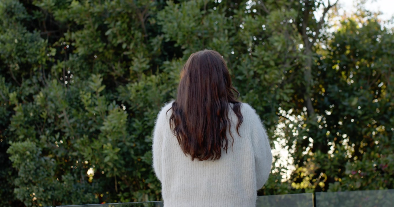 Light breeze is moving hair as woman is standing at glass railing watching sunlight through pines