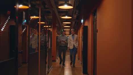 male coworkers going in office hallway male coworkers walking down in hallway together talking about work discussing startup project or business strategy using digital tablet computer marketing company office corridor