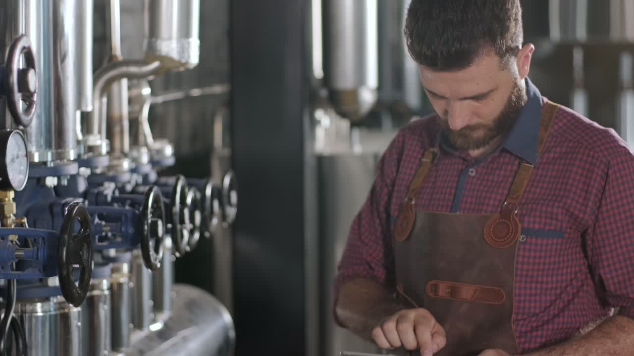 Young male brewer wearing a leather apron supervise the process of beer fermentation at a modern brewery factory