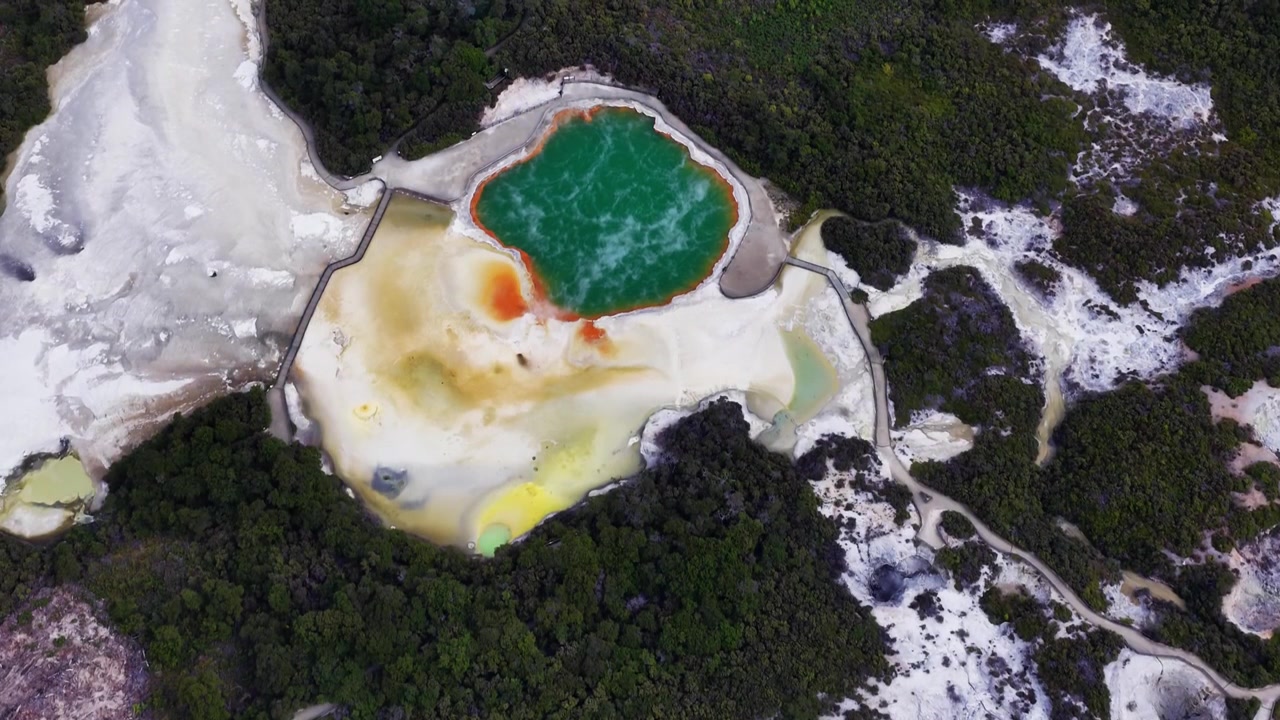 Panoramatický záběr dronu shora na geotermální jezírka Rotorua v Bay of Plenty na Novém Zélandu, zdůrazňující barevné minerální usazeniny, pářící se tyrkysovou vodu, sopečný terén a okolní původní les.