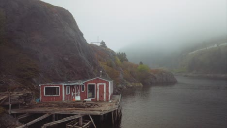 uninhabited wooden home on stilts over calm water nestled against steep cliffs with foggy background