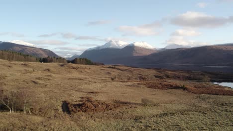 Aerial-drone-footage-rising-above-moorland-and-peat-bog-to-reveal-an-epic-view-of-Glen-Etive-and-Loch-Etive-in-the-Highlands-of-Scotland-with-snow-capped-mountains,-a-forest-and-still-reflective-water