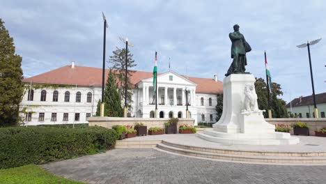 estatua de lajos kossuth frente a la hermosa oficina del alcalde de mako con banderas húngaras colgando en un parque bien cuidado.
