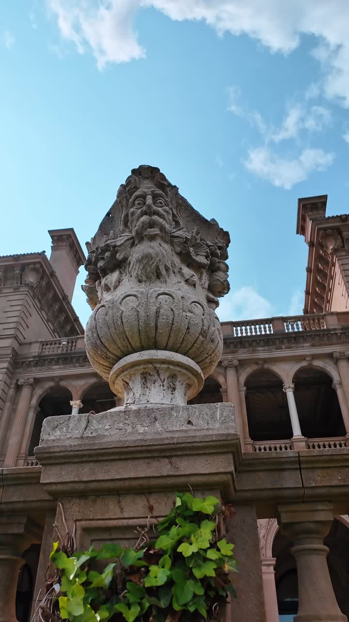 Urna in pietra scolpita con volto su piedistallo da giardino. Vista dal basso di un vaso di pietra logoro con un volto intagliato situato sullo sfondo di una villa