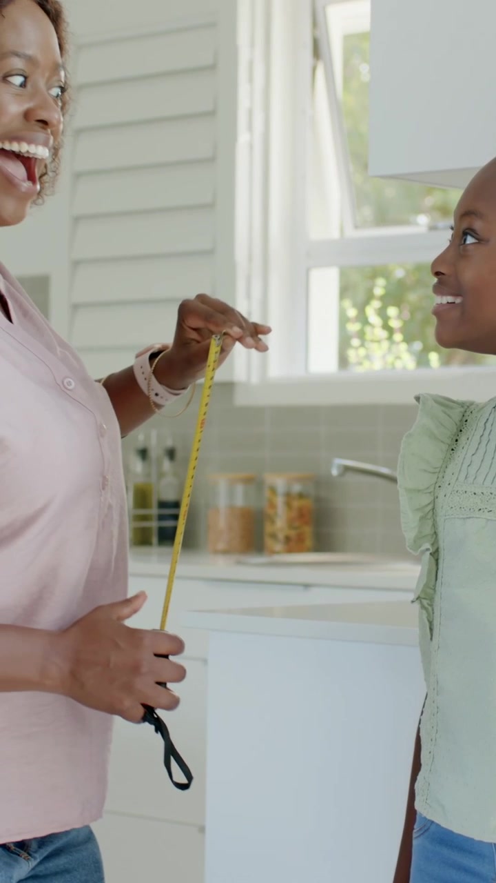 Vertical video: Lifting yellow tape, mother measuring daughter's height in kitchen, wearing green