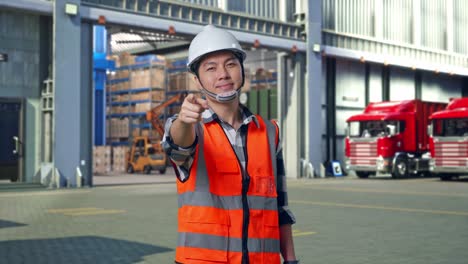 asian male engineer with safety helmet smiling and touching his chest then pointing at you while standing , outside of logistics distributions warehouse