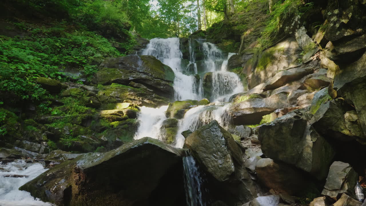 un par de viajeros con mochilas escalando rocas hasta una hermosa cascada de video de 4k y 10 bits