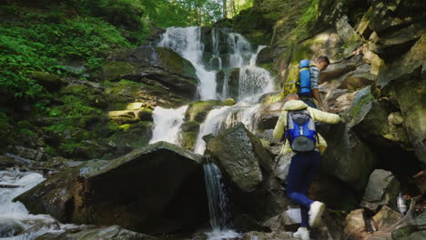 un par de viajeros con mochilas escalando rocas hasta una hermosa cascada de video de 4k y 10 bits