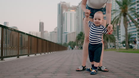 happy cute baby learns to walk and takes first steps in the city under the control of his mom holding baby's hands in the background of the city