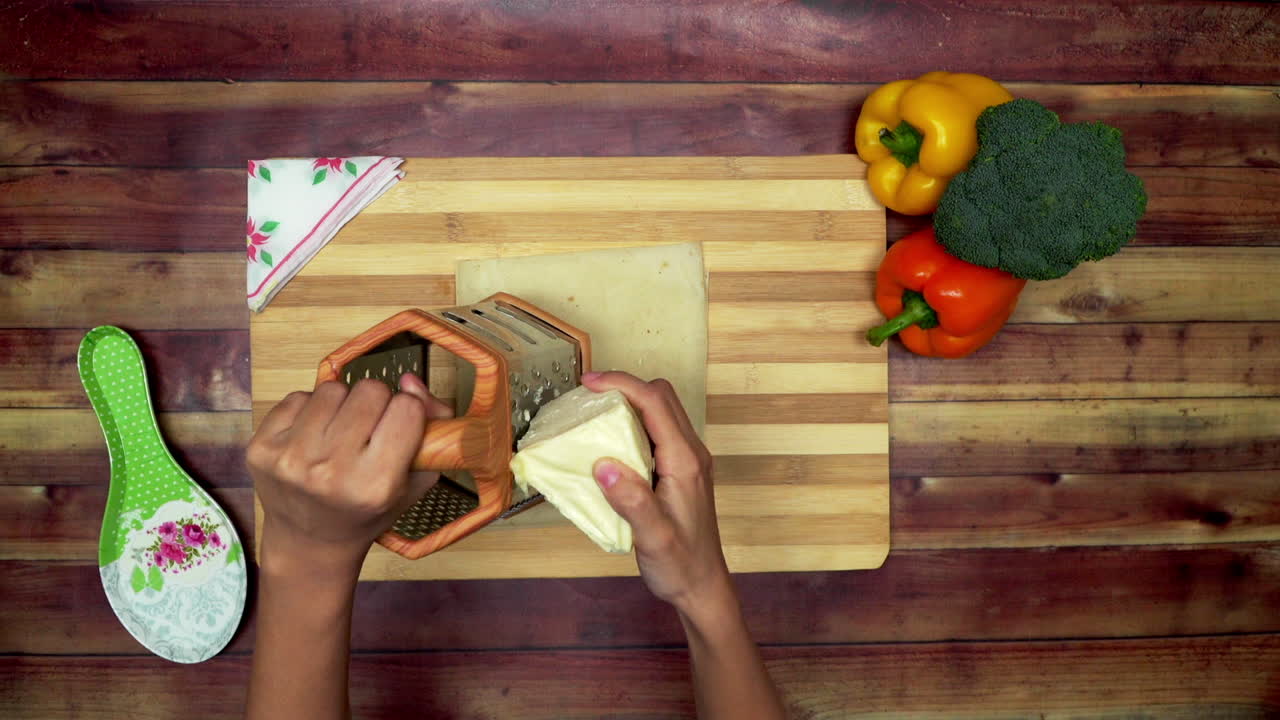 una vista superior de la rotación de la rebanada de queso por un rastrillo, dos grandes capsicums amarillos y rojos, rastrillo y un brócoli verde en la mesa
