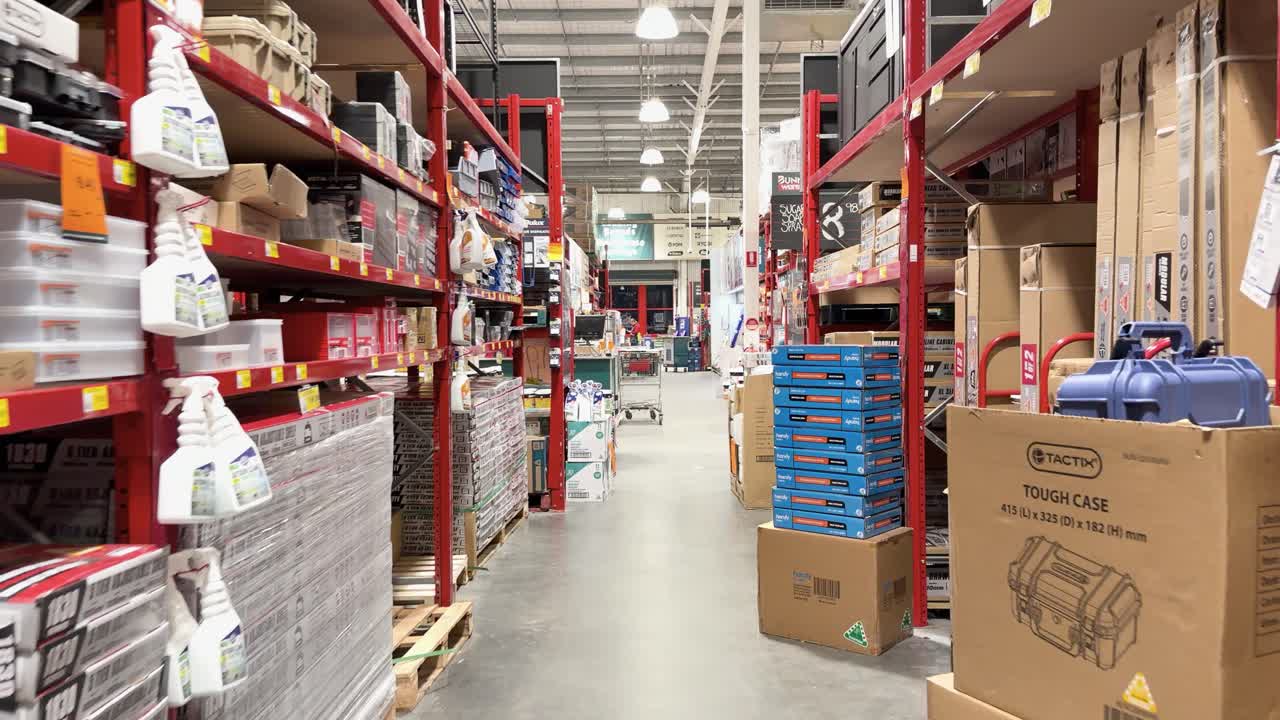 Camera moves through hardware store aisle lined with tiles, paint, tools, and bright lighting