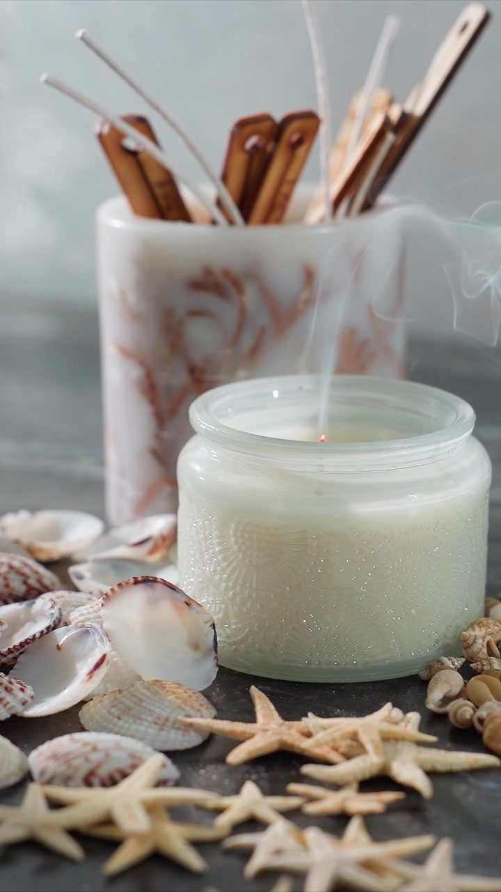 Gros plan sur une bougie allumée dans un verre entourée de coquillages et d'étoiles de mer avec le mouvement de la mèche au fil du temps. La scène comprend des éléments décoratifs comme des bâtons en bois derrière un plus grand récipient, maintenant.