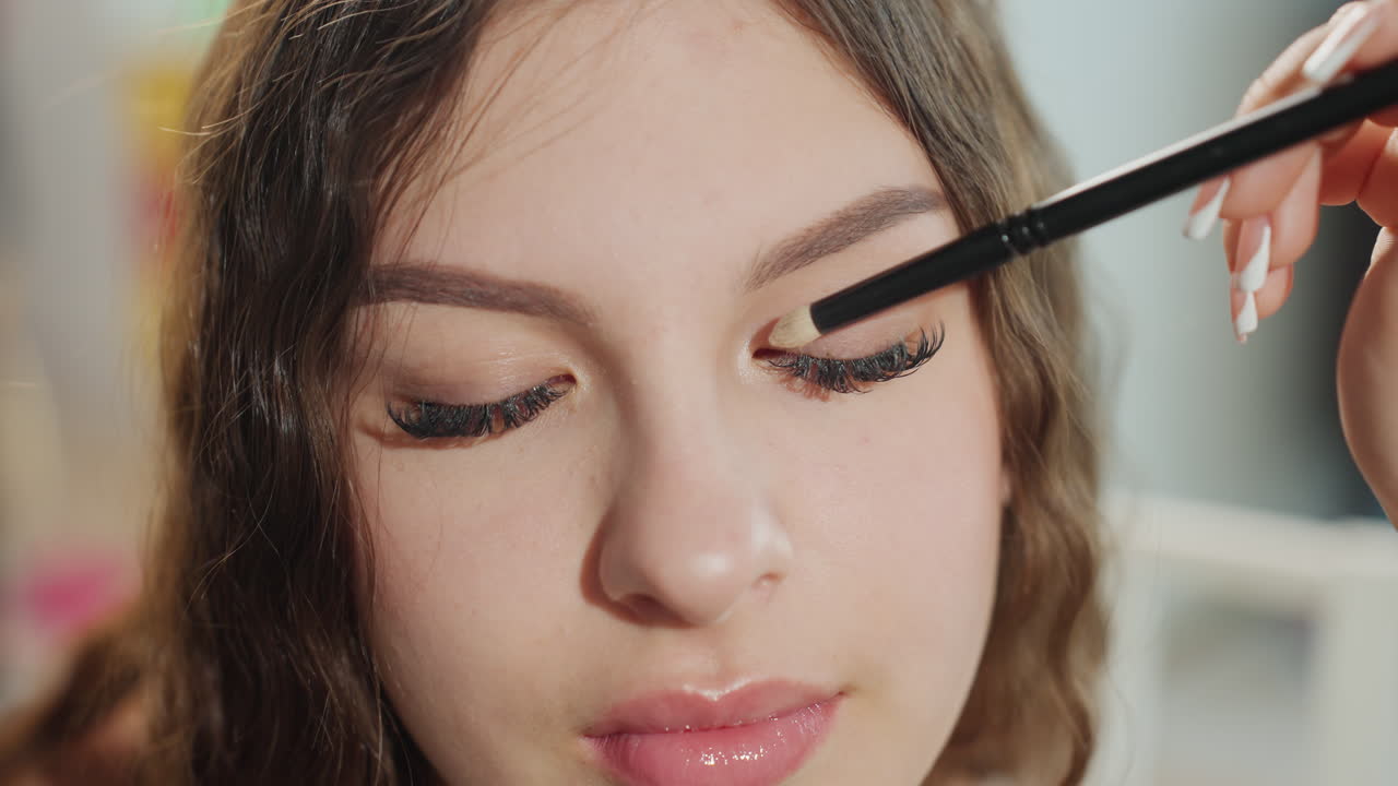 Young Woman Eyes Closed While Brush Applies Eyeshadow, Precise Crease Blending, Long Lashes, Glossy Lips, Natural Skin Finish, Studio Lighting, Model Receiving Professional Makeup