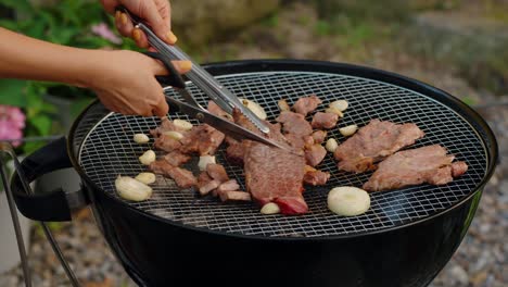 a woman's hands use kitchen shears and tongs to cut grilled beef and cook with garlic and onions during a summer garden barbecue party, showcasing a korean dining style