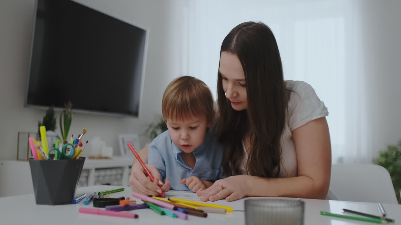 una familia de un niño y una joven madre sentados en la mesa dibuja en papel con lápices de colores. desarrollo de la creatividad en los niños. interior blanco limpio