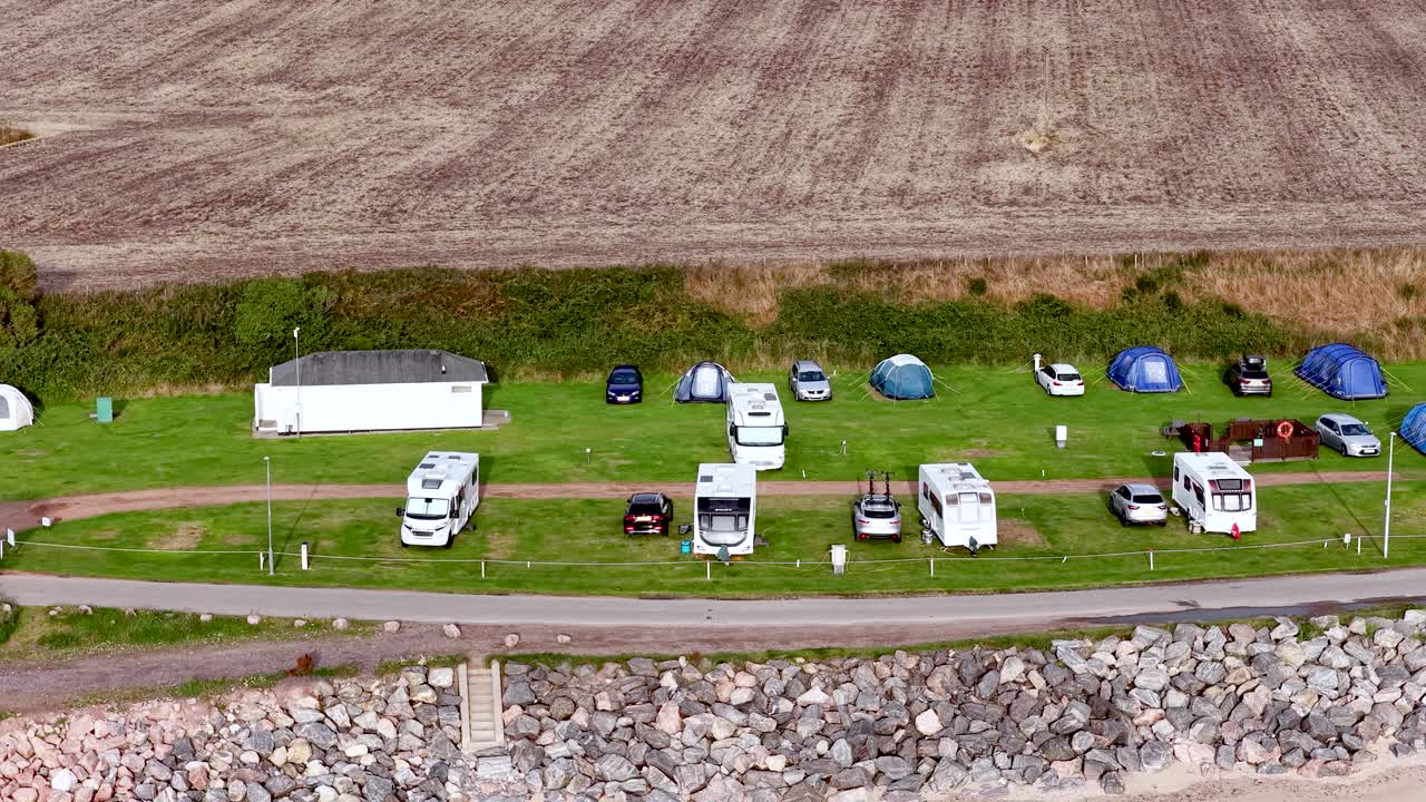 Drone camera moves above seaside campsite with caravans, tents, vehicles, and grassy field in daylight