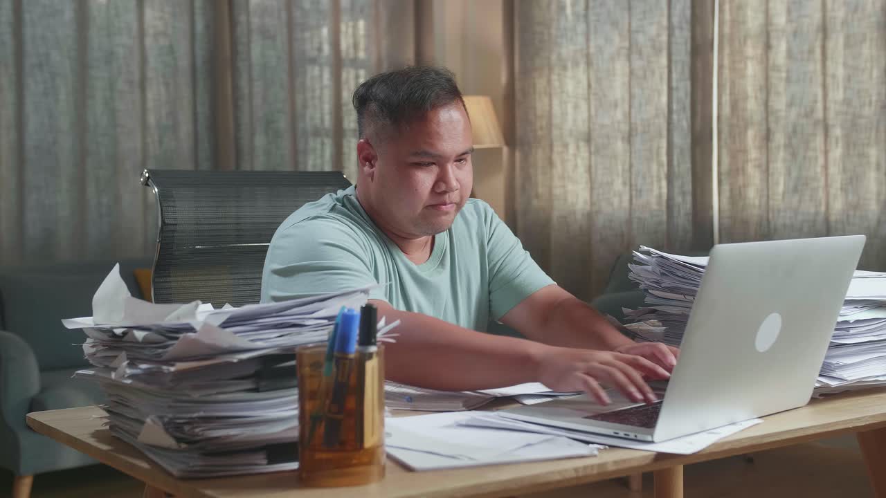 Fat Asian Man Using A Laptop Then Taking Note While Working With Documents At The Office