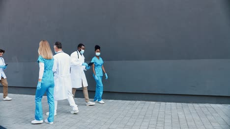 Mixed-raced-male-and-female-doctors-in-white-and-blue-gowns-and-medical-masks-walking-outdoors