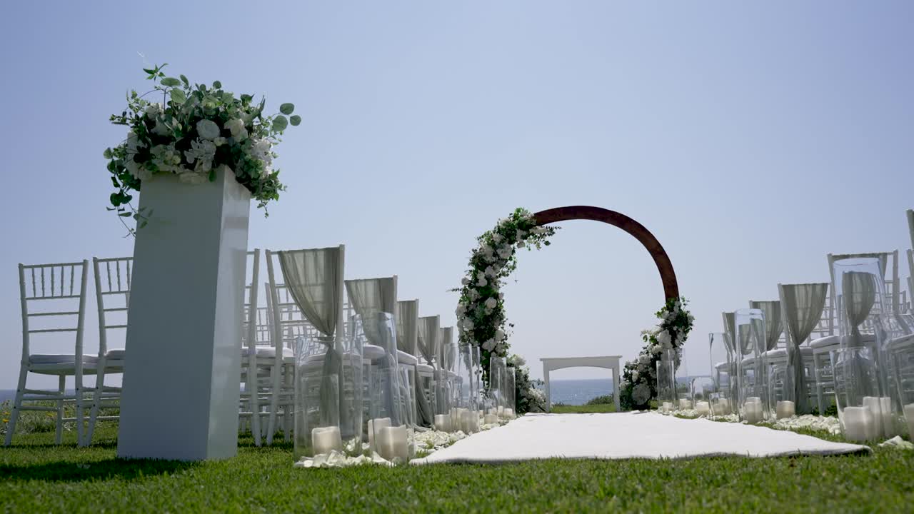 Sedie bianche fiancheggiano un sentiero che conduce a un arco decorato con fiori. Il sole splende alto nel cielo. L'erba verde circonda l'area, creando una splendida cornice per un matrimonio.