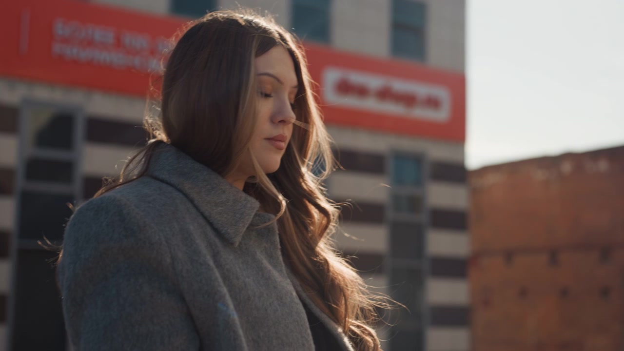 Bella donna bianca su un tetto che compone con una tastiera portatile, indossa un cappotto grigio con lunghi capelli castani, sistema un piccolo controller vicino a un camino, edifici industriali in mattoni sullo sfondo, retroilluminazione calda.