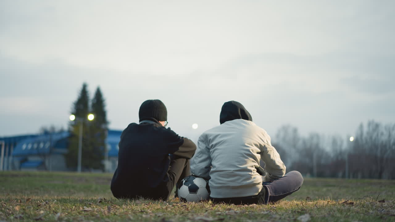 Two boys are seated on a grassy field with their backs to the camera, one in a gray jacket resting his left hand on a soccer ball, with a blur background