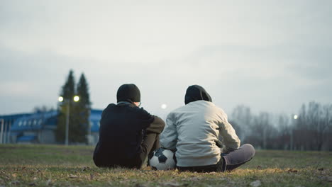 two boys are seated on a grassy field with their backs to the camera, one in a gray jacket resting his left hand on a soccer ball, with a blur background