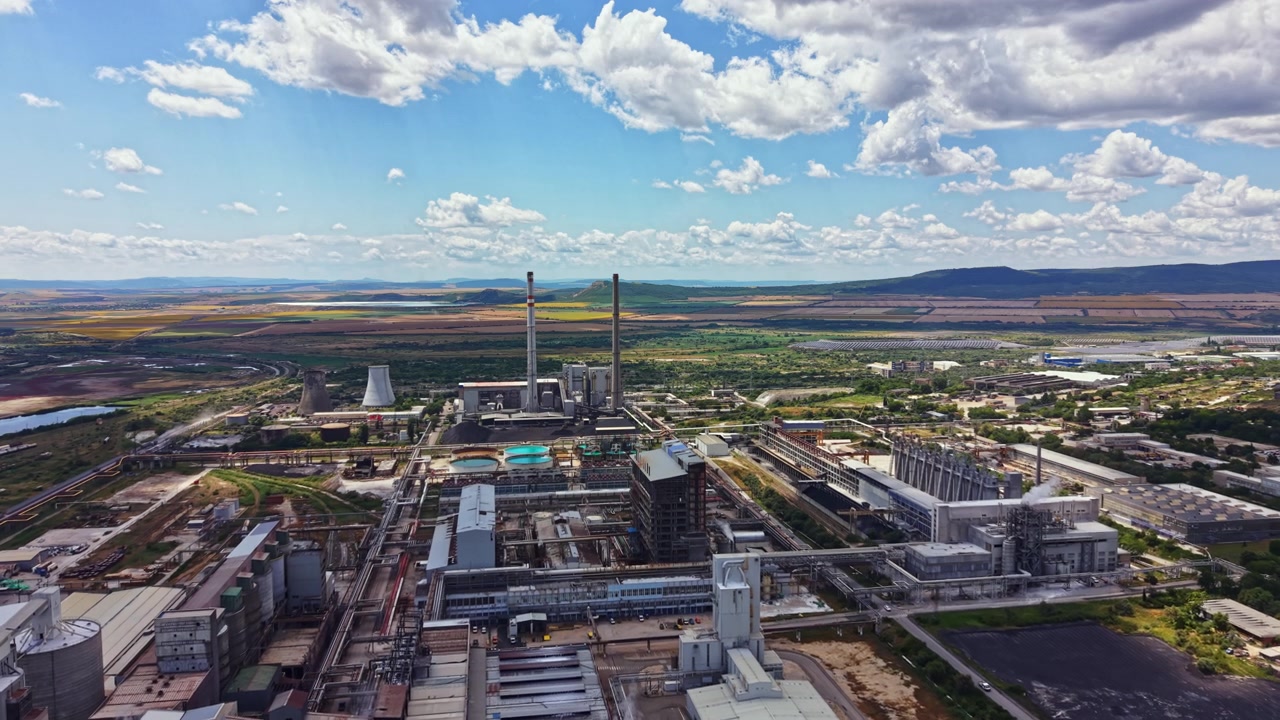 Vista di un grande complesso industriale con torri, edifici e campi verdi sullo sfondo in una giornata di sole con nuvole nel cielo.