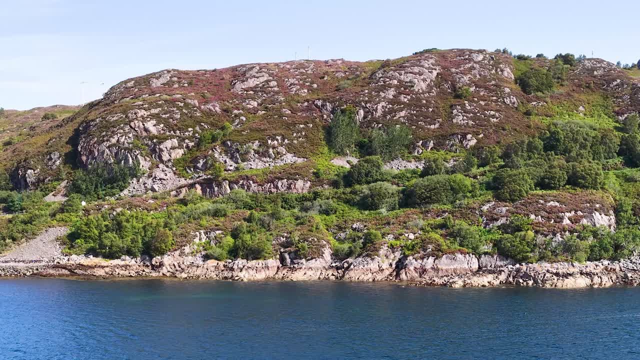 Drone pans along rugged Fife coastline, revealing rocky hills, green vegetation, and calm blue water
