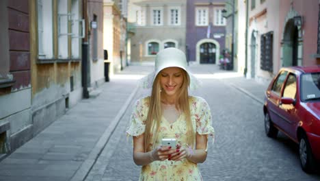 woman texting on street. cheerful woman in white hat walking on old town street and messaging with smartphone