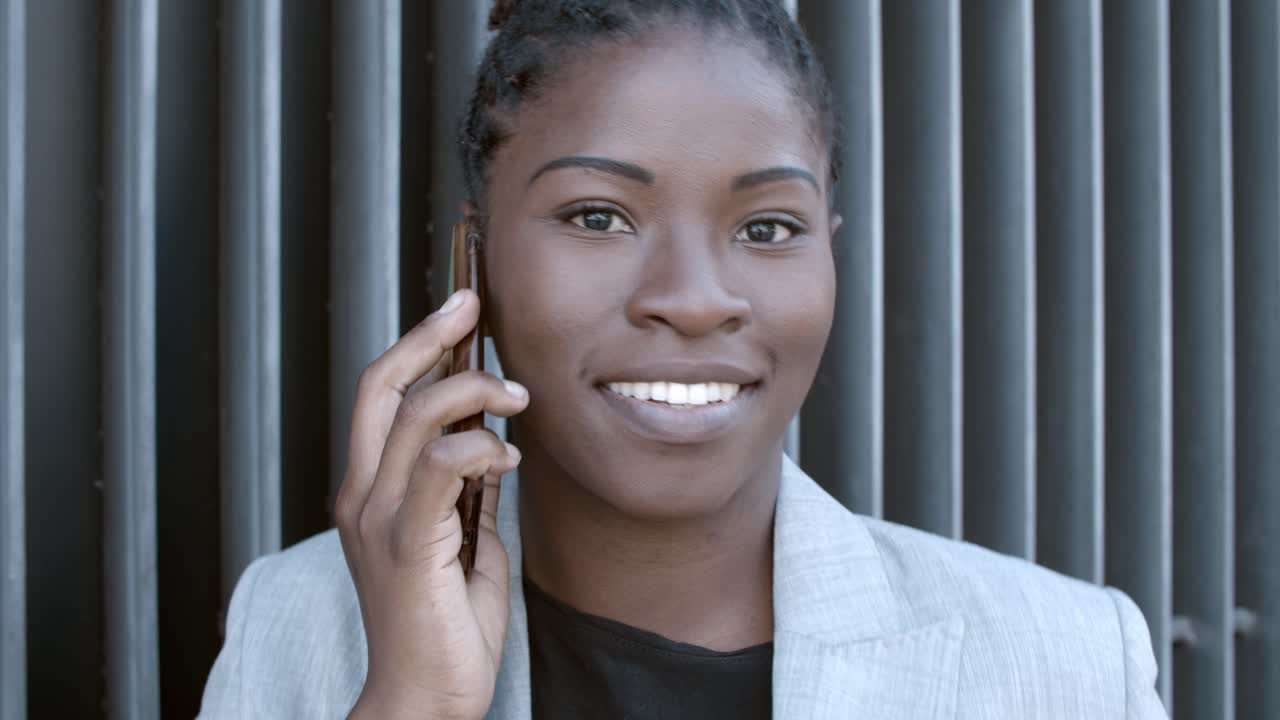 Close up shot of a smiling African-American businesswoman talking on mobile phone and glancing at camera outside