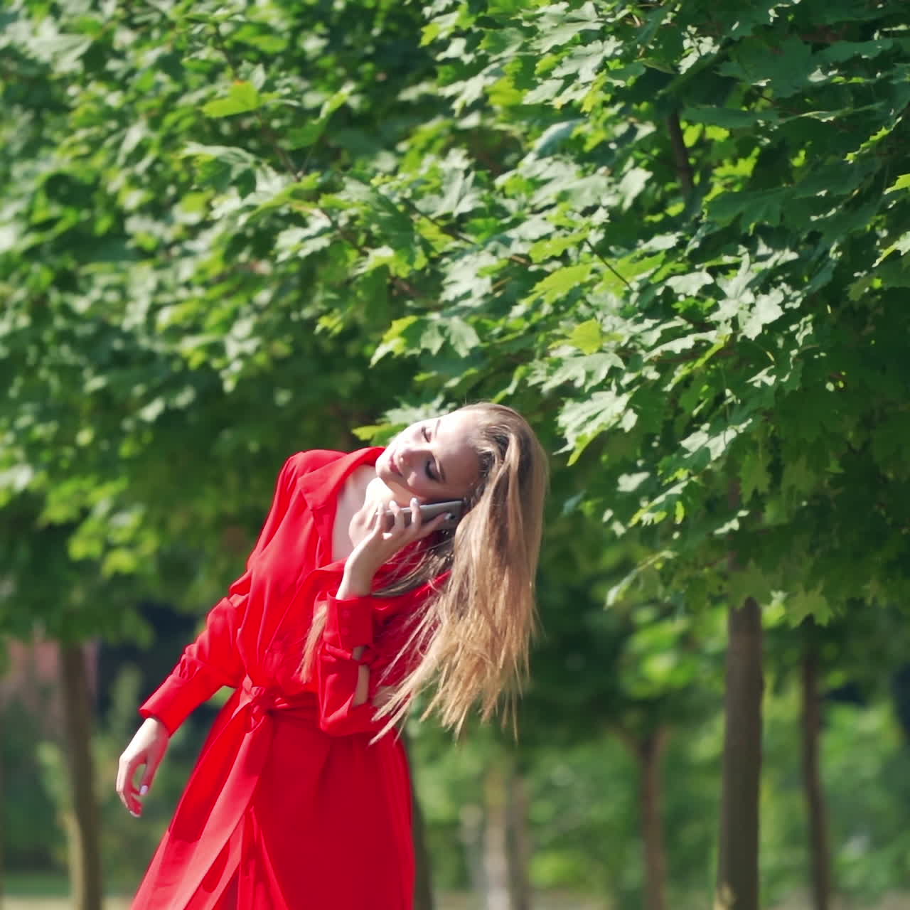 Young woman in beautiful red dress. Lovely female talking on the phone on green trees background. Attractive lady outdoors. Slow motion.