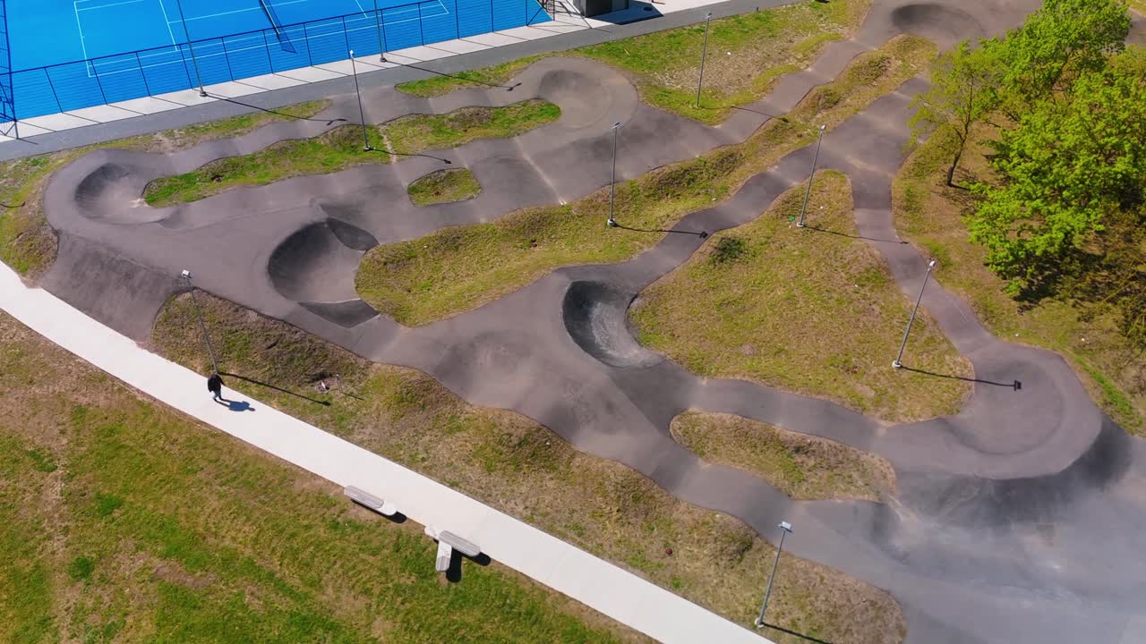 Aerial view of a curvy, paved bike and skate track in a park, surrounded by greenery and adjacent to a tennis court. Ideal for outdoor sports, recreation, and urban park themes.