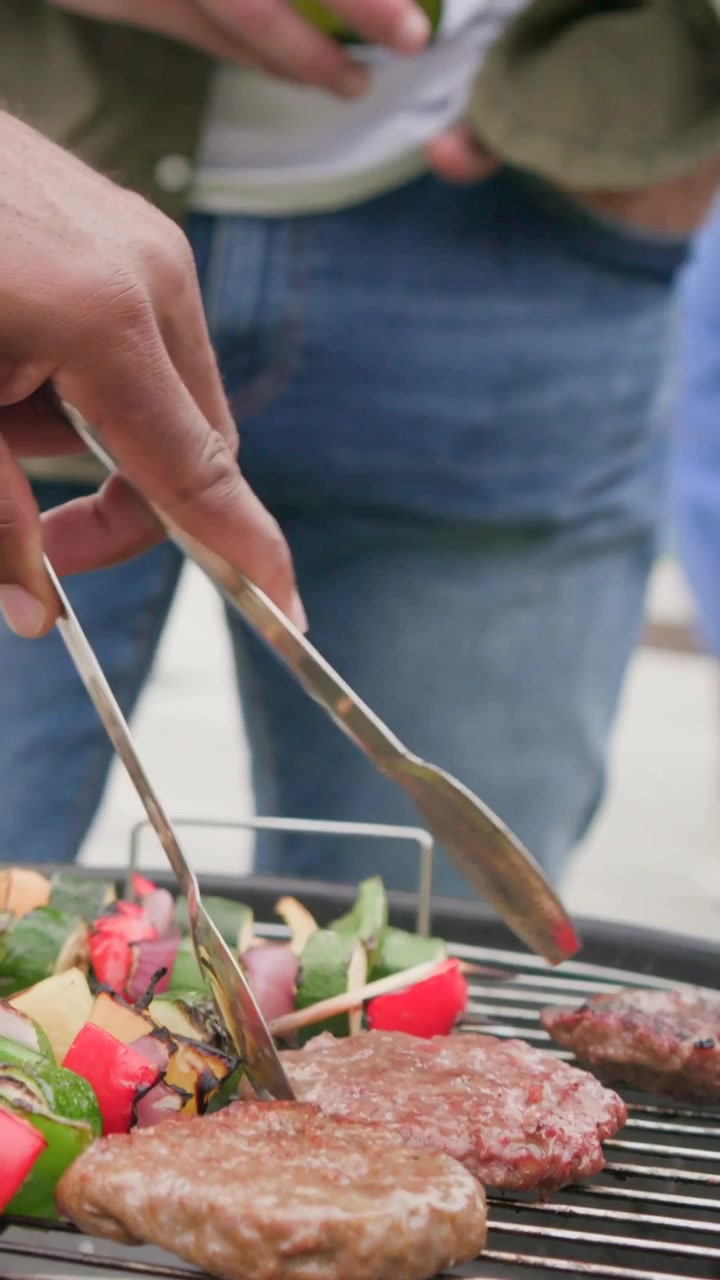 Vertical video: Tongs entering frame, man flipping burgers and skewers on kettle on deck for picnic