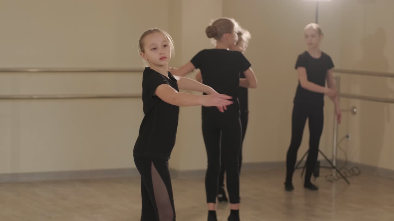 Um grupo de jovens estudantes de balé, vestindo roupas de dança pretas, praticando posições em um espaçoso estúdio de balé com piso de madeira e barras fixadas na parede. Expressões concentradas e movimentos sincronizados