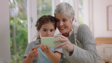 grandmother and little girl using smartphone watching online entertainment eating cookies in kitchen happy granddaughter enjoying sharing weekend with granny browsing internet on mobile phone