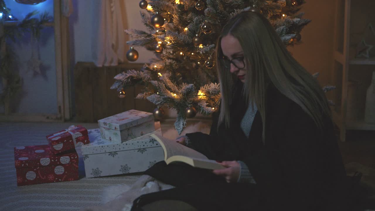 A woman wearing glasses and a cozy sweater sits on the floor reading a book beside a beautifully decorated Christmas tree. Wrapped gifts and glowing lights create a peaceful holiday atmosphere
