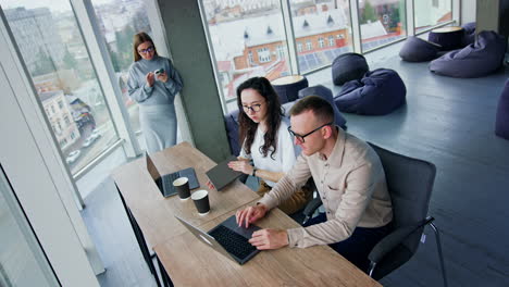 male and female workmates work at laptop at desk, searching information. woman standing at backdrop looks at her phone. high angle view.