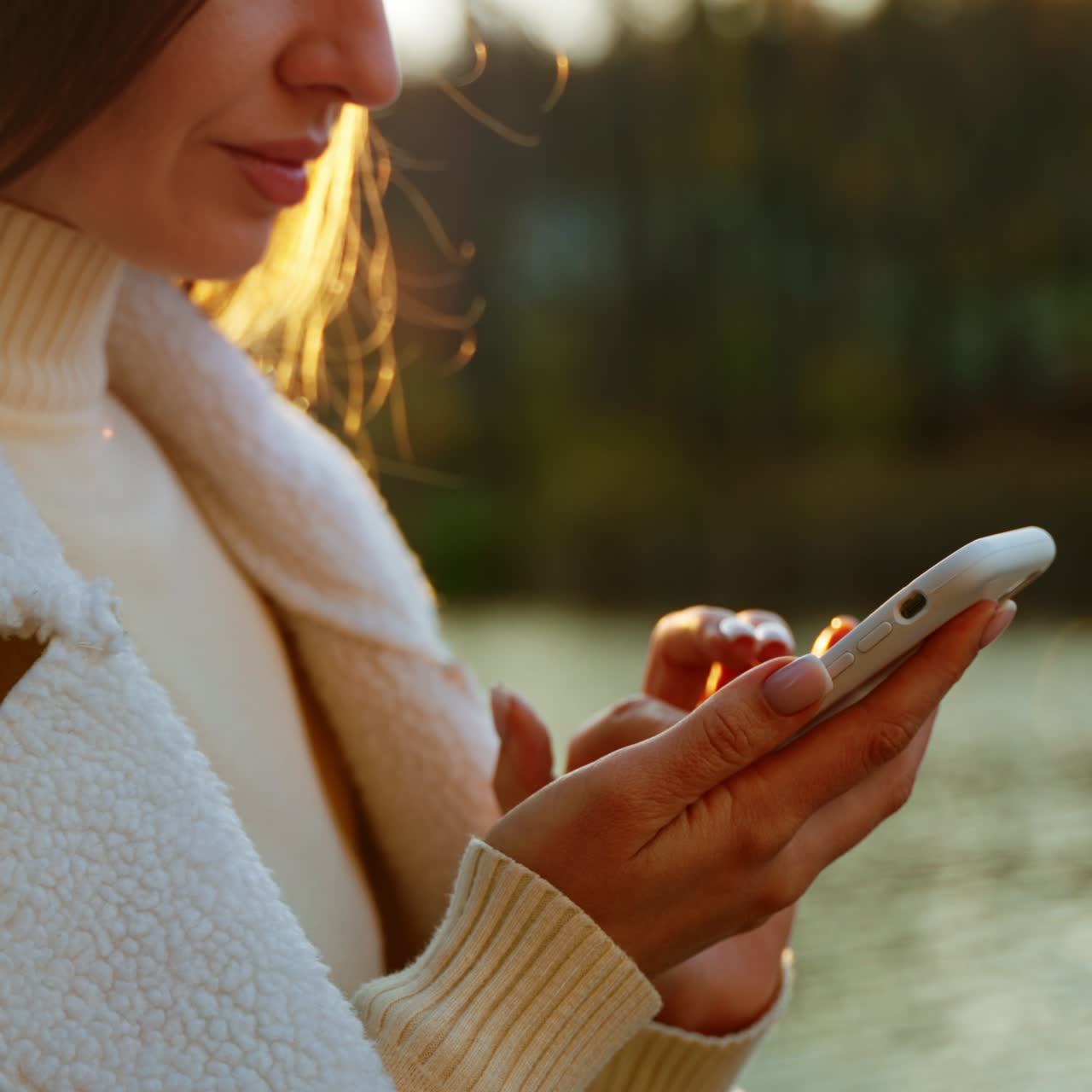 Caucasian dark-haired woman in white jacket using her smartphone outdoors. Rays of sun at blurred backdrop