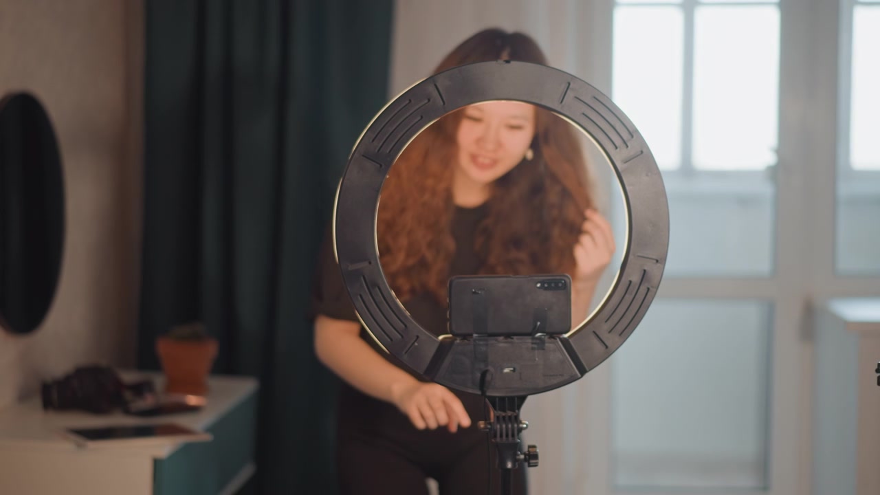 Young person with long hair stands behind circular ring light, holding smartphone mount while making careful adjustments and looking through ring, creating ongoing setup movement in bright indoor room