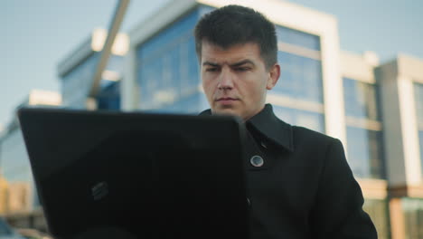 businessman in black coat intensely focused on laptop screen while seated outdoors in urban environment, background features modern office building