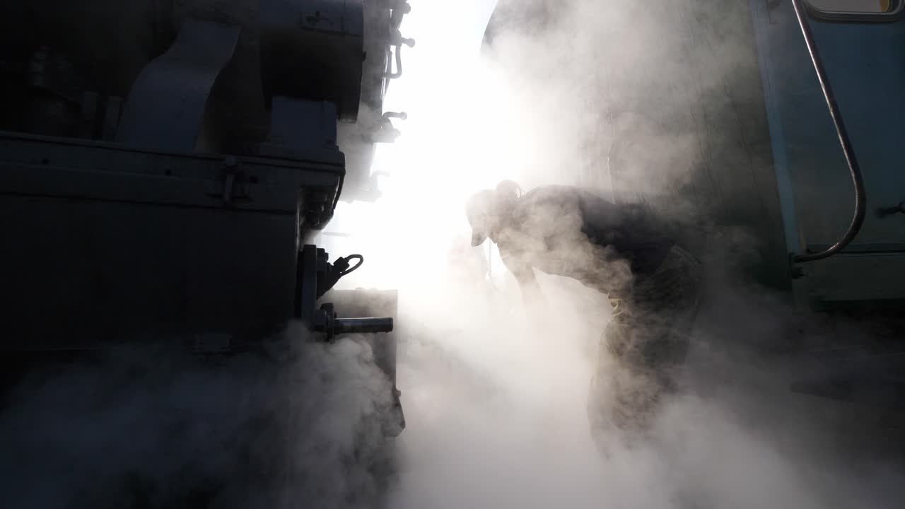 Atmospheric shot of a railroad engineer connecting a historic steam engine to a train. White steam billows around the worker, evoking nostalgia and the golden age of travel