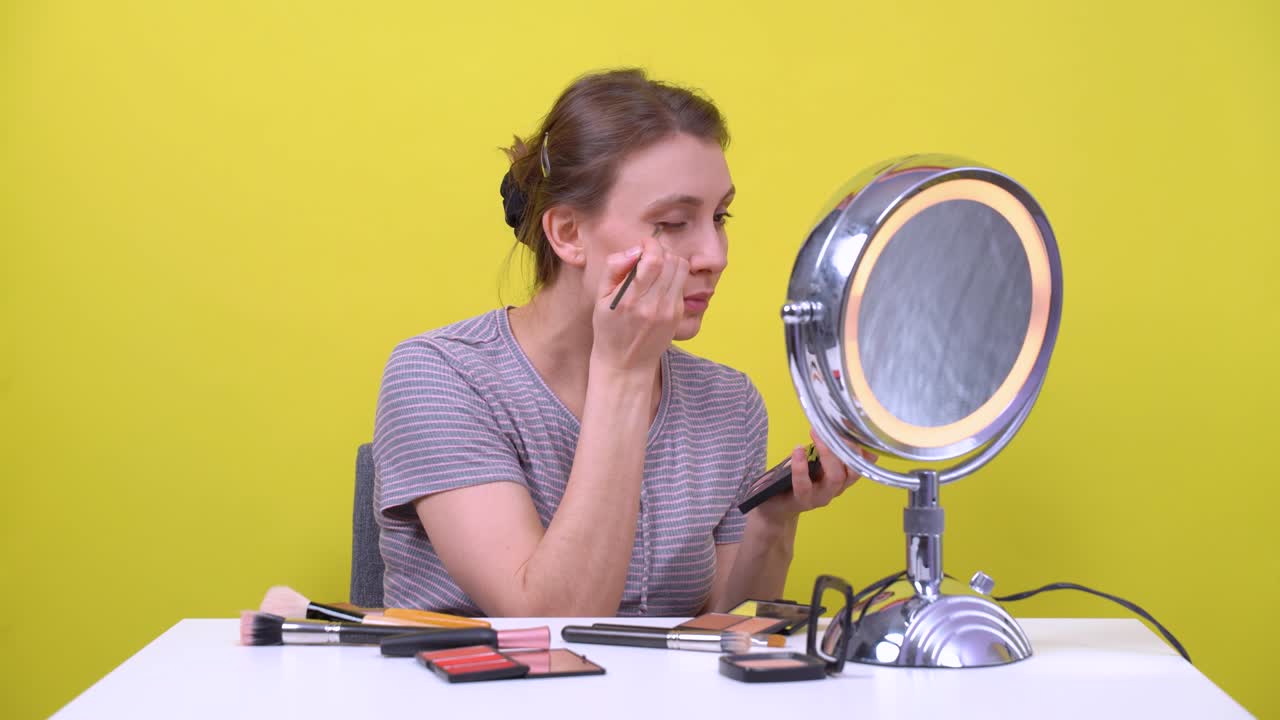 joven mujer atractiva haciendo maquillaje frente al espejo redondo de doble superficie sobre un fondo amarillo brillante. blogger aplicando sombras para los ojos con un pincel cosmético, otros productos de belleza están en la mesa blanca.
