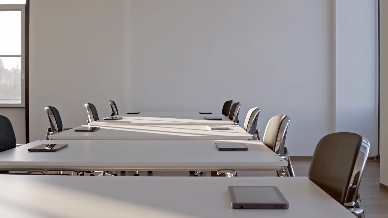 Long low angle view of empty corporate meeting room with parallel white desks, closed tablets near front edges, paired ergonomic chairs, chrome frames, neutral walls, daylight shadows and calm